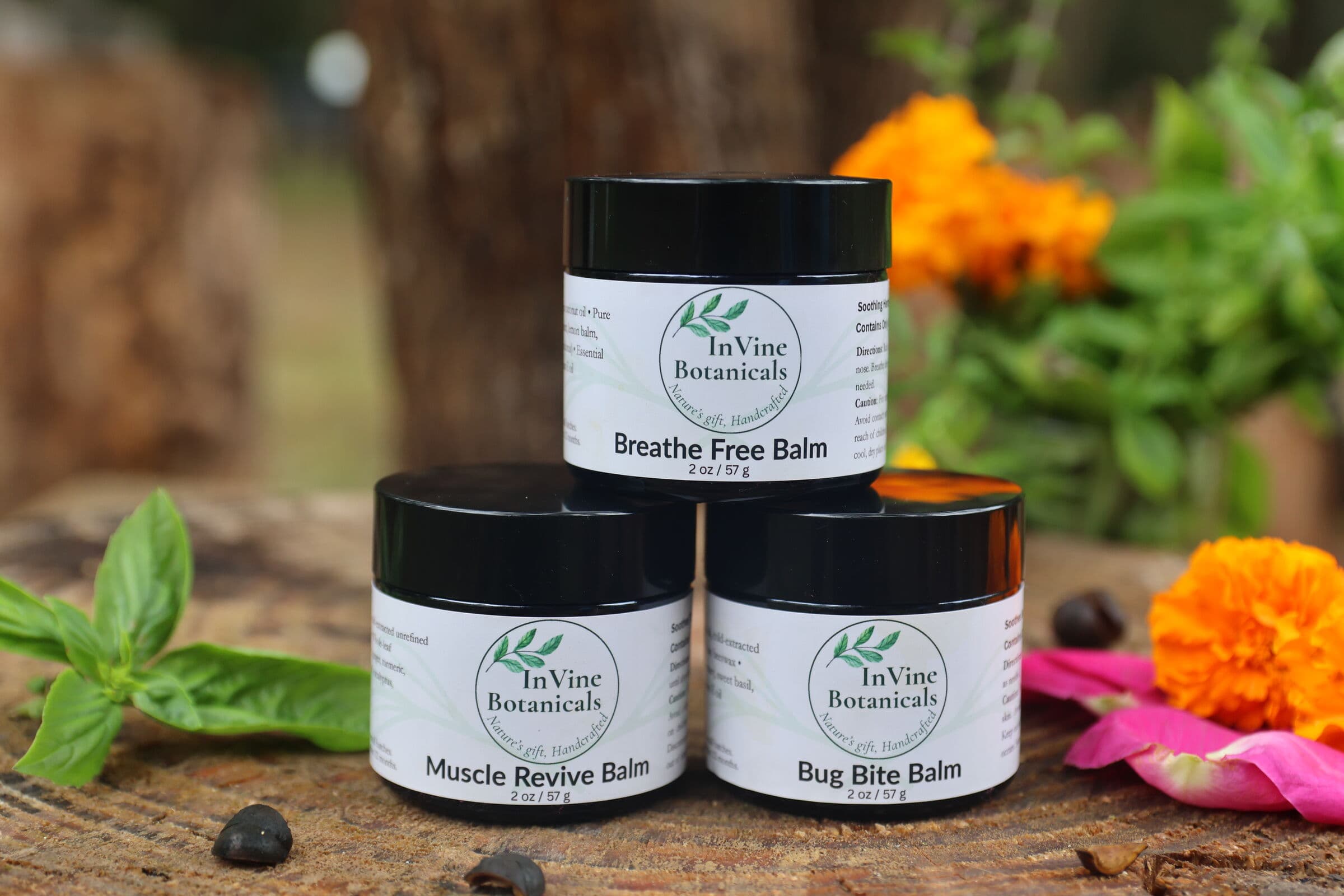 InVine Botanicals Nature's Finest Trio — Bug Bite, Breathe Free, and Muscle Revive whole-herb balms stacked outdoors with fresh basil and marigolds, a non-toxic Mother's Day gift